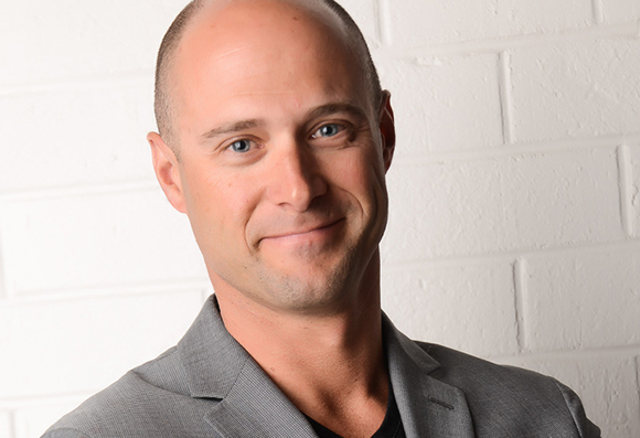 A bald man with blue eyes smiling in a grey blazer against a white brick wall.