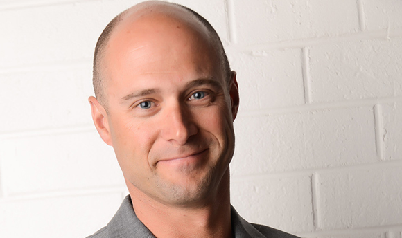 A bald man with blue eyes smiling at the camera in front of a white brick wall.