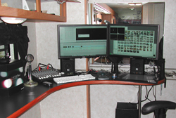 Dual computer monitors, a mixing board, a keyboard, and speakers on a curved black desk.