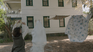 A person in a grey vest hangs white and floral sheets on a clothesline in front of a large white house.