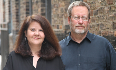 A woman with long brown hair and a man with glasses and a beard standing in front of a brick wall.