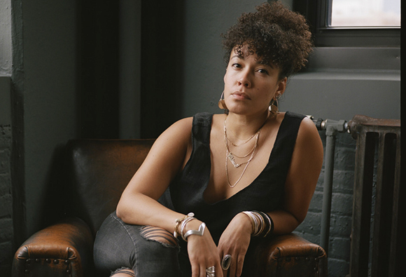 A woman with curly hair and silver jewelry sits in a brown leather armchair against a dark wall.