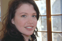 A young woman with dark wavy hair and light eyes smiling in front of a window.