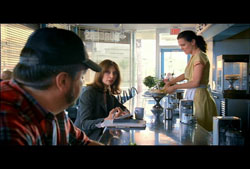 A woman in a dark jacket sits at a counter while a waitress in a yellow dress prepares food in the background.