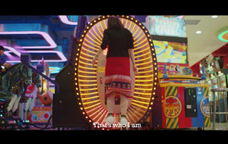 A person in a black shirt and red skirt stands before a large, glowing circular light machine in a colorful arcade.