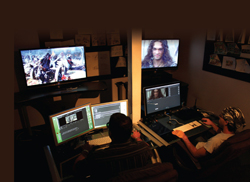 A person sits at a desk with four computer monitors displaying various video content in a dimly lit room.