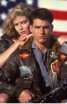 A man in a flight jacket and a woman posing together in front of a large American flag.