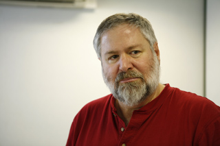 A man with graying hair and a full beard wearing a red henley shirt looking slightly to the side.