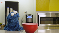 A blue furry puppet sits on a counter next to a large red bowl in a kitchen.