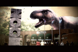 A massive dinosaur head rises behind a building with a tall stone sign that reads ZOO.