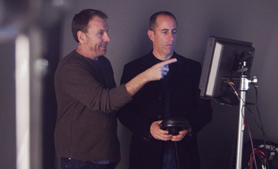 A man in a brown sweater points at a computer monitor while another man in a black jacket watches.