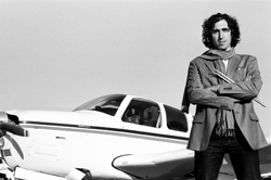 A man with curly hair wearing a blazer and scarf stands with arms crossed in front of a small propeller plane.