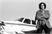 A person with dark hair stands with arms crossed in front of a small propeller plane.