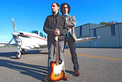 Two men stand on an airfield with a small white propeller plane and hangars under a clear blue sky.