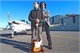 Two people stand on an airfield with a small white airplane in the background under a clear blue sky.