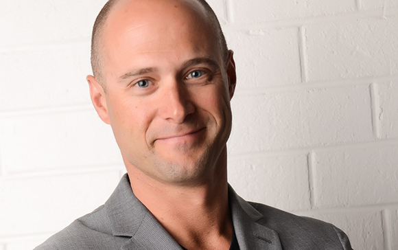 A smiling bald man wearing a grey blazer against a white brick wall.