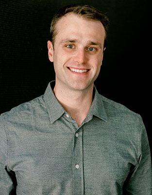A smiling man with short brown hair wearing a grey button-down shirt against a black background.