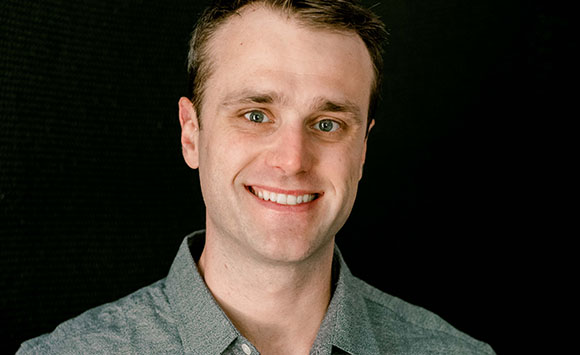 A smiling man with short brown hair wearing a grey button-down shirt against a black background.
