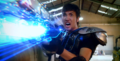 A man in black armor fires a bright blue energy beam from a futuristic weapon in a warehouse.