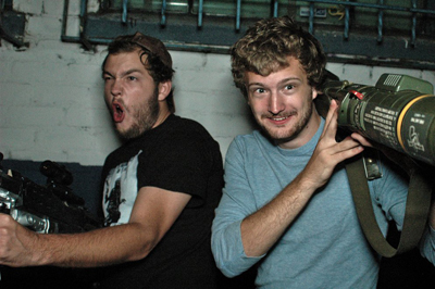 Two men pose indoors; one makes a surprised face while the other holds a large telephoto lens.