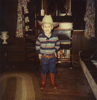 A young child wearing a white cowboy hat, striped shirt, blue pants, and brown cowboy boots.