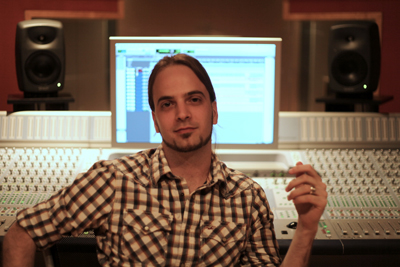 A man in a plaid shirt sits before a large mixing console and computer monitor in a studio.