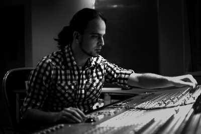 A man in a plaid shirt adjusts controls on a large professional audio mixing console in a dark studio.