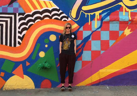 A person in dark clothing stands before a large, vibrant, geometric abstract mural.