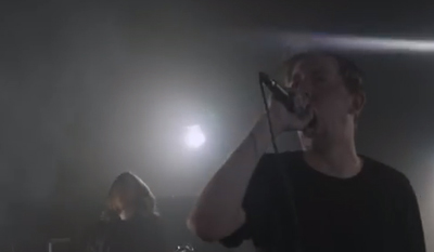 A person in a black shirt sings into a microphone under a bright spotlight in a dark, smoky room.