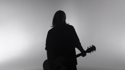 Silhouette of a person with long hair holding an electric guitar against a light gray background.
