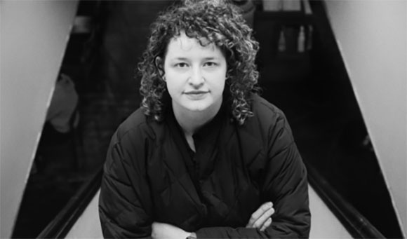 Black and white portrait of a person with curly hair wearing a dark jacket, looking at the camera.