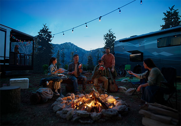 A group of people sit around a campfire at night near RVs and trees under a twilight sky.