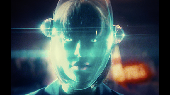 A person's face illuminated by a glowing, translucent futuristic helmet against a dark, blurred background.