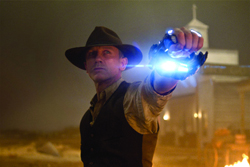 A man in a cowboy hat and vest points a futuristic glowing blue device toward the camera.
