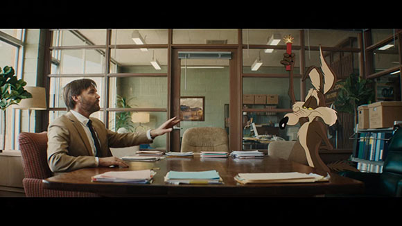 A man in a tan suit gestures toward a cartoon coyote holding a lit sparkler in an office.