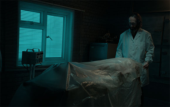 A man in a white lab coat looks down at a body covered by a translucent plastic sheet in a dark room.