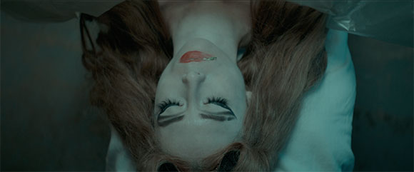 A person with long hair and heavy eye makeup lies on a white surface, viewed from an upside-down angle.