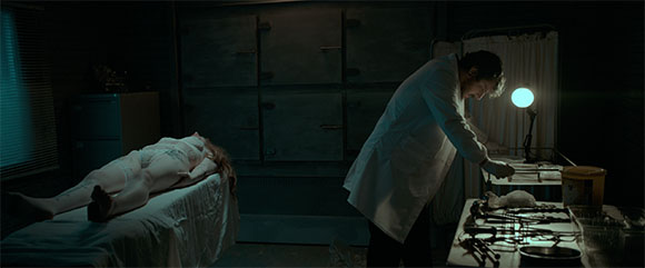 A person in a white lab coat leans over a table next to a person lying on an examination table in a dark room.