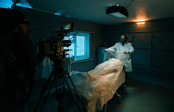 A camera operator films a person in a white lab coat covering a body with a plastic sheet in a dark room.