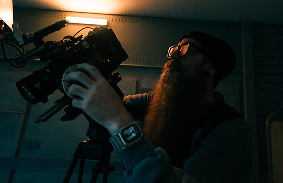 A bearded man in a beanie adjusts a large professional film camera on a tripod in low light.