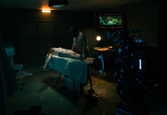 A film camera captures a scene of a man standing over a person lying on a medical table in a dark room.