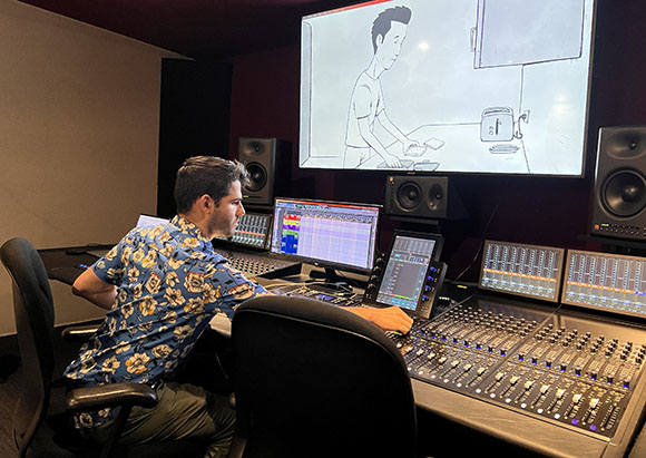 A man in a floral shirt sits at a large mixing desk with multiple monitors in a dark studio.