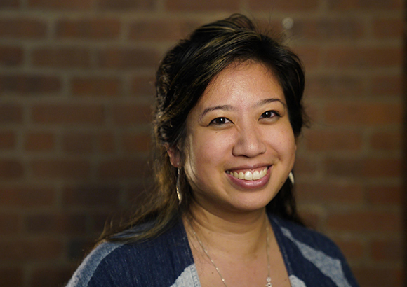 A smiling woman with dark hair wearing a blue patterned top against a brick background.
