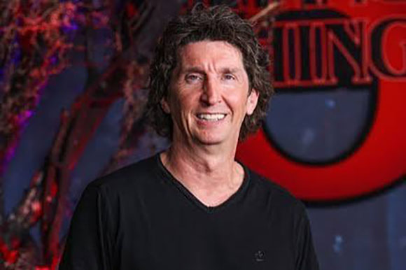 A man with wavy brown hair smiling in a black v-neck shirt against a dark, colorful background.