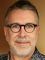 Close-up portrait of a man with short grey hair, blue-rimmed glasses, and a grey beard against a tan background.