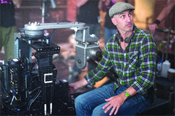 A man in a green plaid shirt and flat cap sits beside a large black robotic camera rig.