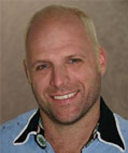 A smiling man with light hair and stubble wearing a light blue collared shirt.