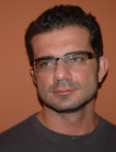 A man with short dark hair and glasses looking toward the camera against an orange background.
