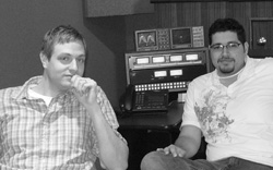 Two men sit in a studio next to a rack of audio recording equipment.