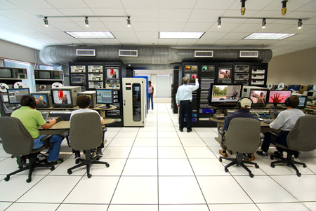 People working at computer workstations in a control room filled with video monitors and technical equipment.